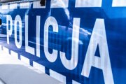 Zdjęcie przedstawiające napis Policja na srebrnych drzwiach radiowozu oznakowanego, koloru niebieskiego.