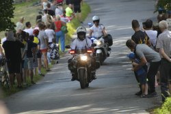 policjanci na motocyklach pilotujący kolarzy na drodze powiatowej, a na poboczach kibice czekający na kolarzy