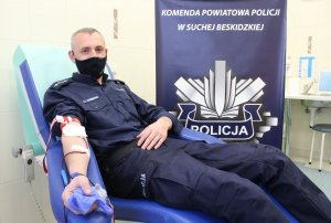 Policjant oddaje krew, w tle baner z napisem Komenda Powiatowa Policji w Suchej Beskidzkiej.