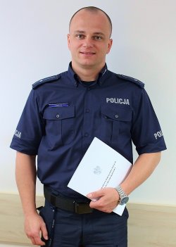 Zdjęcie nowego kierownika dzielnicowych młodszego aspiranta Pawła Bercala trzymającego teczkę z rozkazem personalnym Komendanta Powiatowego Policji w Suchej Beskidzkiej