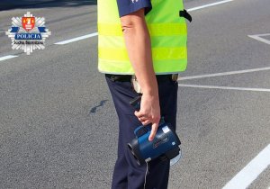 Zdjęcie przedstawia umundurowanego policjanta ruchu drogowego stojącego bokiem i trzymającego w prawej dłoni laserowy miernik prędkości.