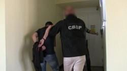 Zdjęcie przedstawiające prowadzonego zatrzymanego skutego kajdankami z tyłu w korytarzu prowadzonego przez policjanta CBŚP a drugi z policjantów otwiera drzwi