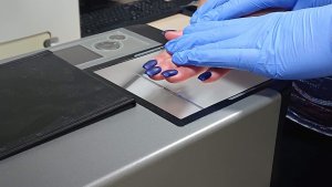 Dłoń damska z pomalowanymi na niebiesko paznokciami na której dwie dłonie ubrane w rękawiczki gumnowe dociskają do urządzenia w tracie pobierania odcisków palców