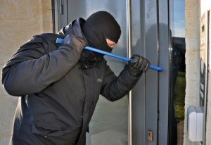 Zdjęcie przedstawia mężczyznę z metalowym łomem koloru niebieskiego przykładanym do drzwi wejściowych koloru szarego i próbującego je otworzyć