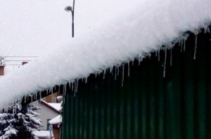 Zdjęcie przedstawiające dach budynku na którym znajduję się duża warstwa śniegu wraz z soplami oraz zieloną ścianę budynku wykonaną z zielonej blachy