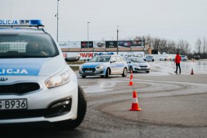 Fotografia przedstawia radiowozy policyjne podczas wykonywania ćwiczenia „slalom” pomiędzy pachołkami. Kilka radiowozów porusza się „jeden za drugim”. W oddali widoczny jest instruktor prowadzący szkolenie ubrany na czerwono.