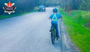 mały chłopiec jadący drogą w kasku i szelkami odblaskowymi i jadący z na przeciwka samochód osobowy