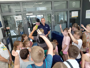 Dzieci w wejściu do komendy trzymające dłonie podniesione i policjant witający uch w wejściu