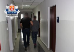 policjant kryminalny trzyma zatrzymanego za lewą rękę i wspólnie idą korytarzem komendy