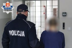 umundurowany policjant trzymający zatrzymanego za lewą rękę i stoją razem przezd kratami pomieszczeń dla zatrzymanych