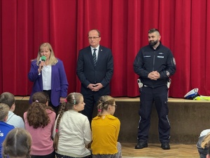 Otwierająca spotkanie pani dyrektor z mikrofonem w ręku i obok stojący sekretarz i suski policjant