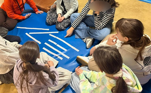 Uczniowie zgromadzeni i siedzący na matach rozwiązują układankę