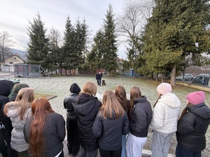 Młodzież ustawiona przed trawnikiem na którym przewodnik psa pokazuje posłuszeństwo podopiecznego