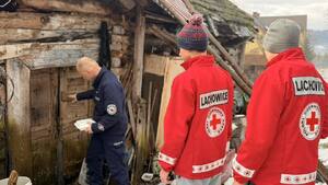 Dzielnicowy wchodzący do bardzo skromnej drewnianej chatki a za nim wolontariusze PCK