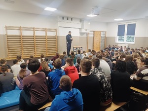 Zebrana grupa dzieci siedząca na materacach w dużej sali słucha wykładu policjanta