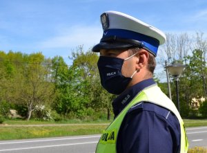 Policjant ruchu drogowego (biała czapka, odblaskowa kamizelka), w stopniu sierżanta sfotografowany od pasa w górę, nadzoruje ruch – obserwuje drogę. W ręce trzyma miernik prędkości. W tle wiosenna, zielona aura.