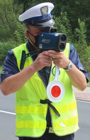 Policjant ruchu drogowego stojący obok drogi i trzymający laserowy miernik prędkości skierowany w stronę jadących samochodów.