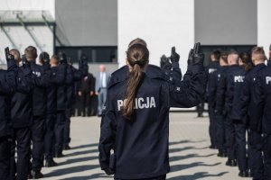 na pierwszym planie stoi tyłem policjantka z uniesioną prawą ręką, w tle po jej obu stronach rzędy policjantów