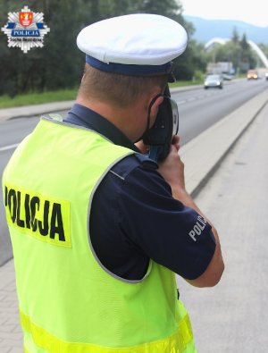 umundurowany policjant stojący obok drogi i mierzący prędkość jadącym samochodom  urządzeniem laserowym