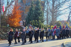 przemarsz orkiestry dętej na ulicy kościelnej w suchej beskidzkiej oraz idące poczty sztandarowe na których przodzie idzie poczet policji