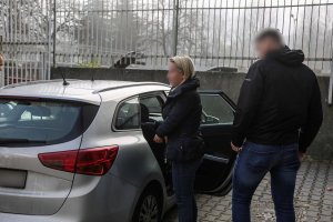 kobieta stojąca przed nieoznakowanym radiowozem z zasłonietą twarzą a za nią stojący nieumundurowany policjant z wydziału kryminalnego