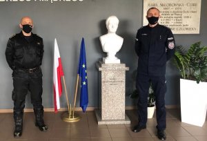 strażak wraz z policjantem stojący przy tablicy patrona szkoły oraz flagi polski i unii europejskiej