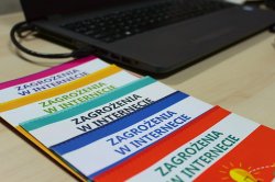 Na biurku ułożone różnokolorowe foldery związane z zagrożeniami w sieci obok których stoi laptop z widoczną klawiaturą