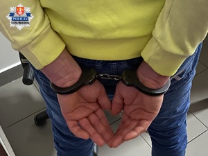 ręce mężczyzny skute kajdankami z tyłu ubranego w żółty sweter. W lewym górnym rogu logo KPP Sucha Beskidzka.