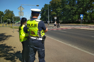 policjantka i policjant stojący przy drodze i obserwujący jadących kierujących oraz pieszą wraz z wózkiem na przejściu dla pieszych