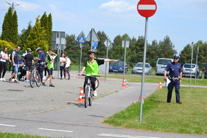 Na miasteczku ruchu drogowego na rowerze jeździ zawodniczka, ocenia ja policjant ruchu drogowego. Przed miasteczkiem stoi grupa oczekujących zawodników.