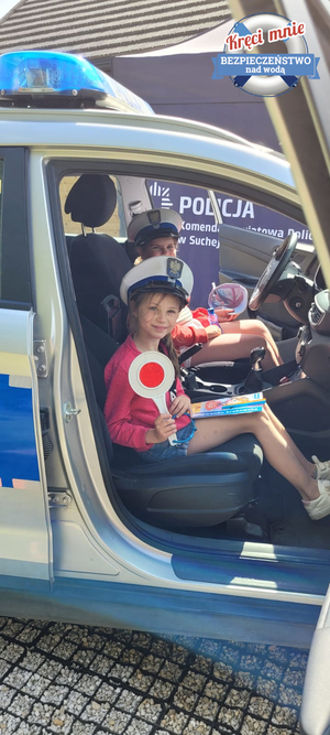 dziewczynka siedzące w radiowozie  z czapką policyjna i tarczą