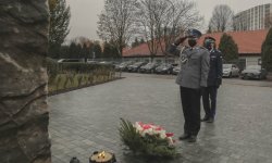 policjant w mundurze galowym salutuje przed pomnikiem przed którym położono wieniec z kwiatami. Za policjantem stoi Komendant Wojewódzki Policji w Krakowie.