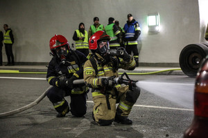 strażacy ubrani w maski tlenowe trzymający węża i lejący wodę  podczas podczas ćwiczeń w tunelu