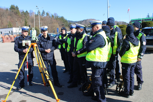Grupa policjantów ruchu drogowego omawiają zasadę pracy urządzenia do laserowego pomiaru punktów przy sporządzaniu planu sytuacyjnego podczas zdarzenia drogowego