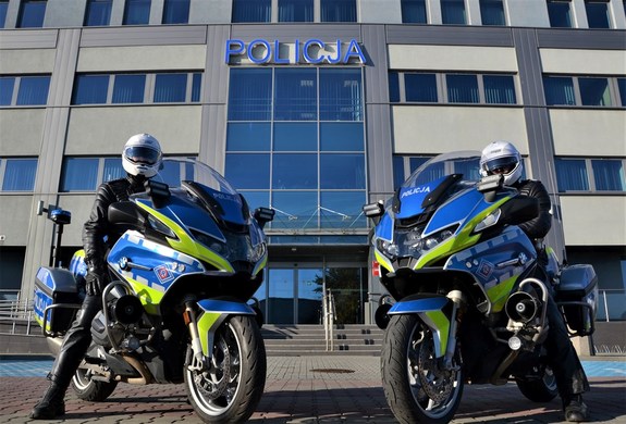 Dwóch umundurowanych policjantów w kaskach, siedzą na policyjnych motocyklach przed budynkiem, na którym widnieje napis Policja