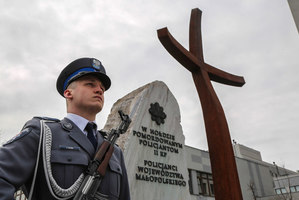 policjant w mundurze galowym a za nim pomnik w hołdzie pomordowanym policjantom 2 rp oraz krzyż