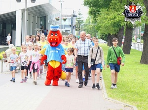 dzieci wraz z rodzicami idą chodnikiem prowadzeni przez suską maskotkę