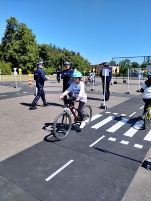 Dziewczynka na rowerze jedzie po miasteczku ruchu drogowego, sędziują policjanci ruchu drogowego