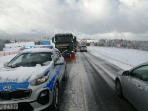 Zdjęcie przedstawiające drogę pokrytą śniegiem po której na jednym pasie znajduje się oznakowany radiowóz z włączonymi światłami a za nim stojący pług śnieżny. Po drugiej stronie po pokrytej częściowo śniegiem poruszają się samochody osobowe.