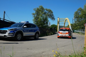 stojący oznakowany radiowóz i jadący biało pomarańczowy samochód rajdowy na metalowy w kierunku dużego mostu