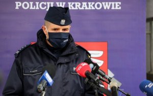 Komendant Wojewódzki Policji w Krakowie stojący na tle baneru komendy przemawia do stojących przed nim mikrofonów różnych przedstawicieli mediów.