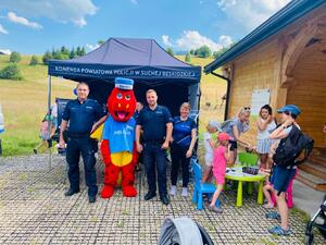 Stoisko Suskiej Policji i pozujący policjanci i pracownica Policji a obok stolik przy którym dzieci rozwiązują krzyżówki