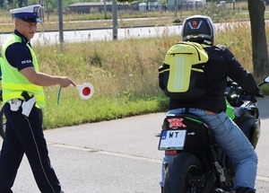 policjant na drodze zatrzymujący jadącego motocyklistę z plecakiem przy użyciu tarczy sygnalizacyjnej