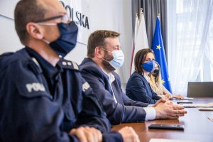 Siedzące przy stole trzy osoby w maseczkach z których pierwszy przy stole siedzi policjant a za nim mężczyzna w garniturze i kobieta