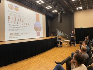 zdjęcie przedstawiające scenę gdzie na ekranie widać plakat debaty oraz siedzących przed scena mieszkańców