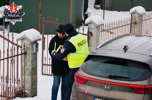 policjant stojący za brązowym samochodem wręcza księdzu opaskę i zakłada na rękę