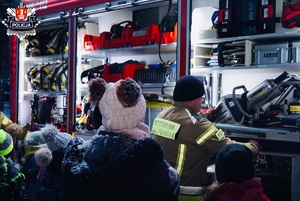 dzieci w czapach i kurtkach zimowych stojące przy beczkowozie strażackim oglądają szczypce hydrauliczne prezentowane przez strażaka